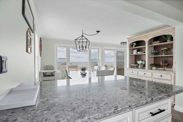 A kitchen with a marble countertop, white cabinetry, and a view of the beach. - Duckling Crossing | Beach House on the Sand (Westbrook)