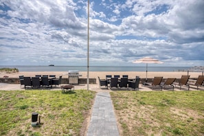 An outdoor seating area with tables and chairs facing a beach, a grill, and a pathway leading to the beach.