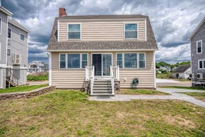 A two-story house with a front porch, a stone retaining wall, and a grassy yard. - Duckling Crossing | Beach House on the Sand (Westbrook)