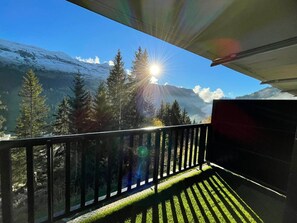 A balcony with a view of snow-capped mountains, trees, and the sun shining through the foliage.