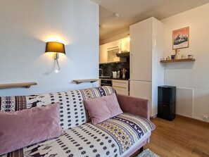 A living room with a sofa, a wall-mounted lamp, a kitchen area with cabinets and appliances, and a framed picture on the wall.