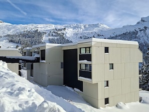 A modern, multi-story building with a flat roof, surrounded by snow-covered mountains and trees.