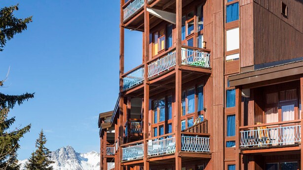 A multi-story wooden building with balconies, large windows, and a mountainous backdrop.