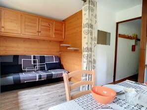 Ett mysigt rum med en väggpanel av trä, en soffa med ett geometriskt mönster på klädseln, en trästol, ett bord med en mönstrad duk och ett fönster med en gardin.