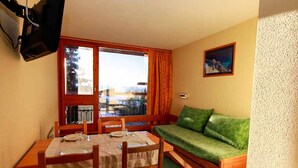 A compact room with a dining table set for two, a green sofa, and a window with curtains. - Ski-to-door studio with sunny balcony (Les Arcs 1800)