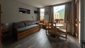 Una habitación acogedora con un sofá de madera, una mesa de comedor con sillas, un ventilador de techo y una ventana con cortinas.