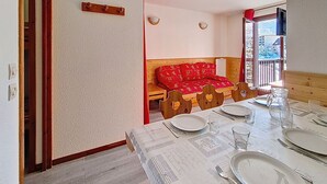 A dining area with a wooden table set for four, a red sofa, and a window with a view of buildings. - Spacious and bright apartment at the foot of the slopes with balcony (Les Menuires)