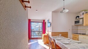 A dining area with a table set for four, wooden chairs, a kitchen with appliances, and a balcony with red curtains. - Spacious and bright apartment at the foot of the slopes with balcony (Les Menuires)