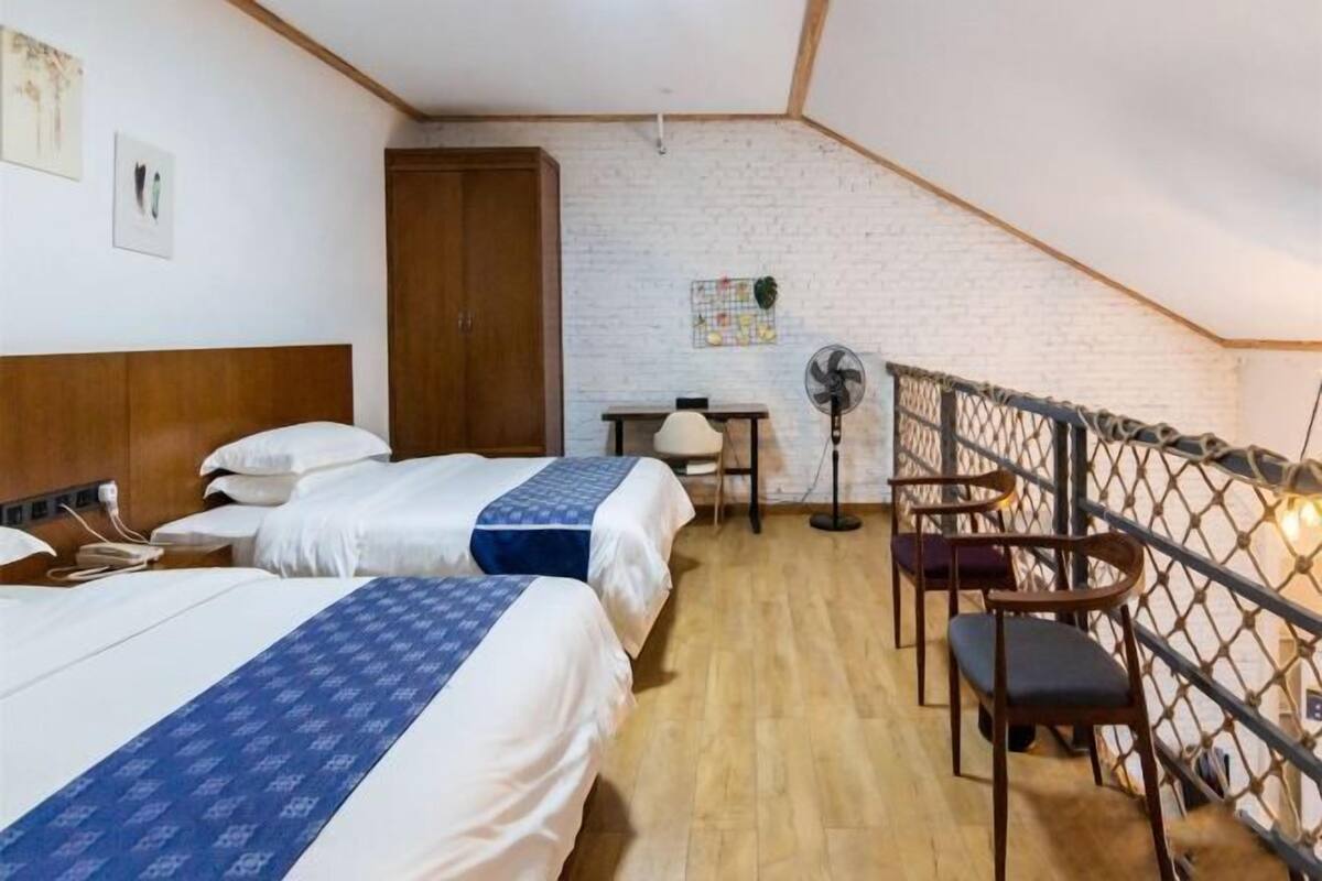 A room with two beds, a wooden headboard, a small table with a fan, and a balcony with chairs.