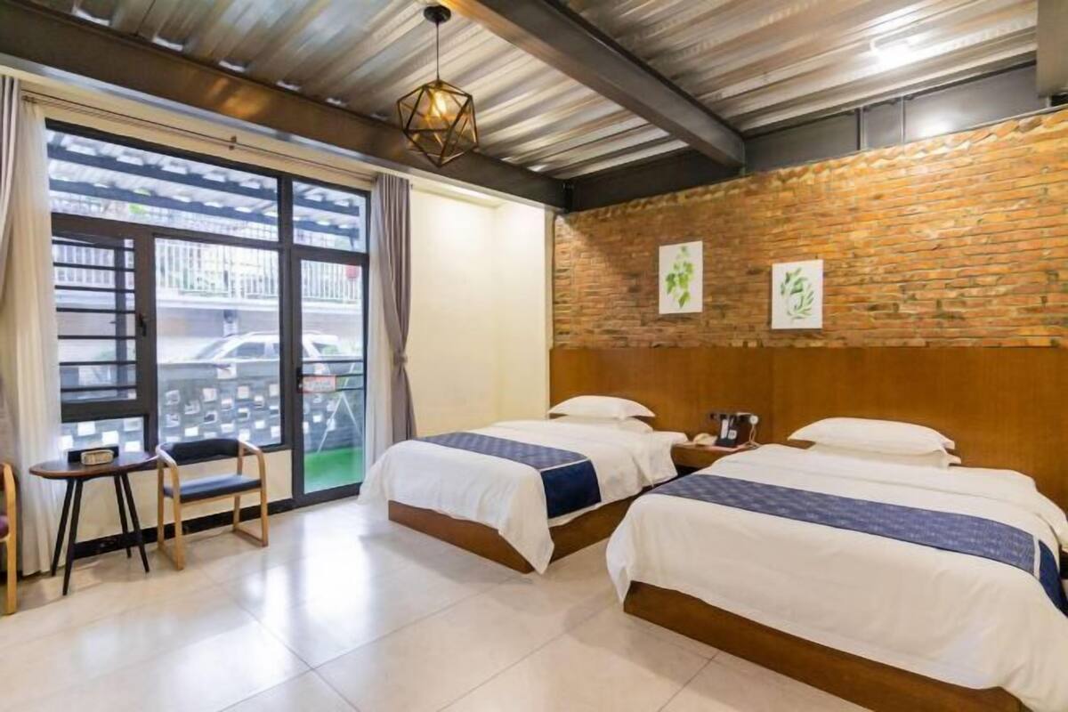 A hotel room with two beds, a small table, and a chair. There is a brick wall and a window with a view of the street.