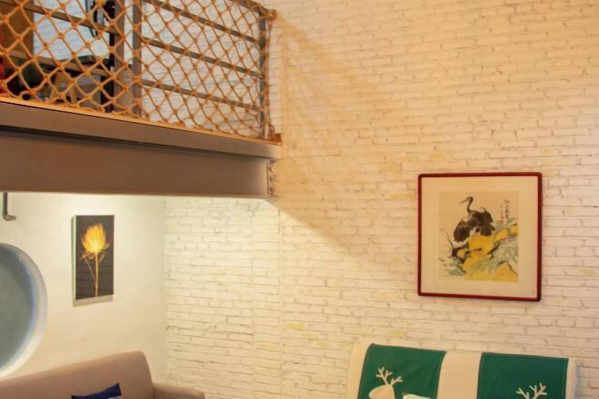 A room with a white brick wall, a round mirror, a green and white striped cushion, and two framed artworks.