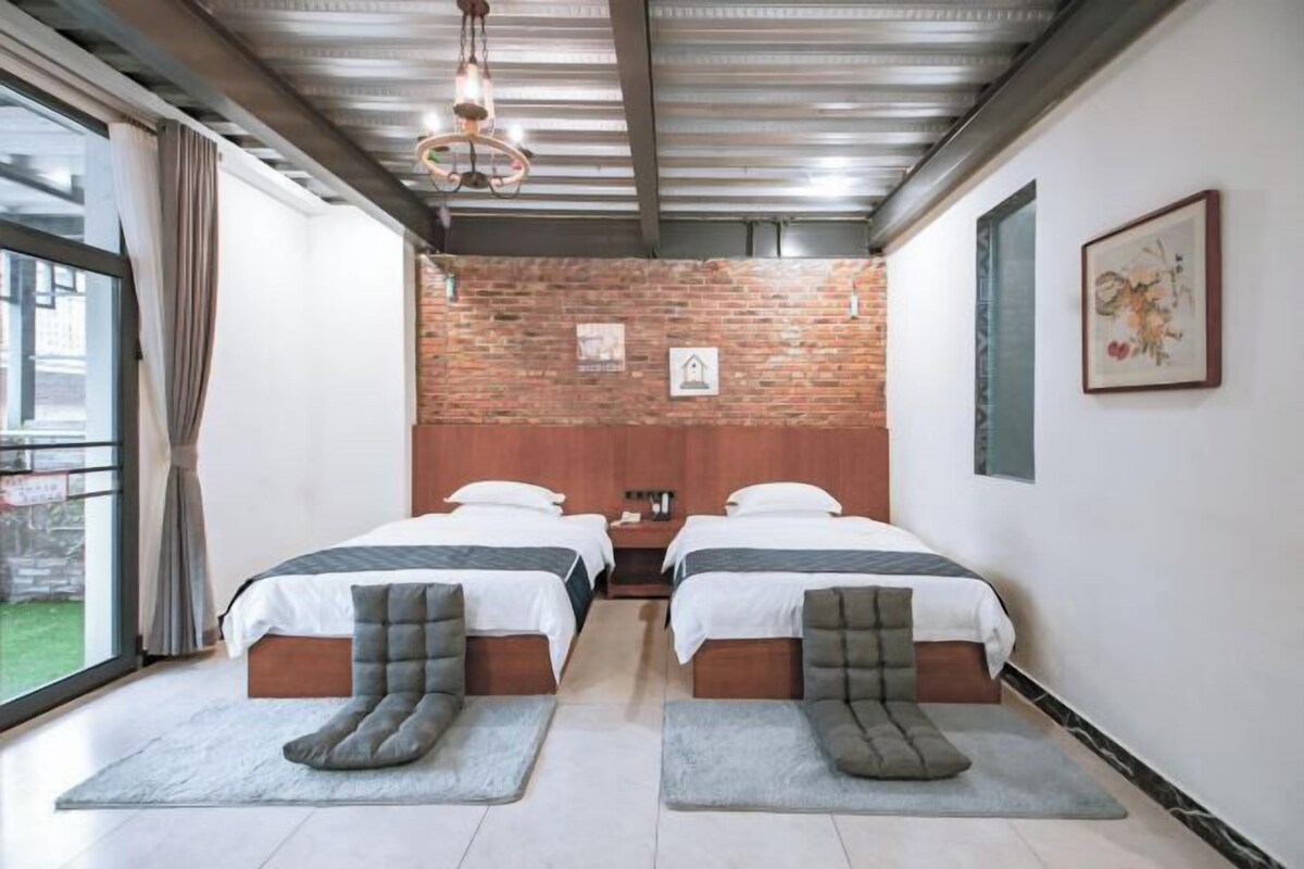 Habitación de hotel con dos camas, una pared de ladrillo, un ventanal grande y un techo con vigas a la vista.