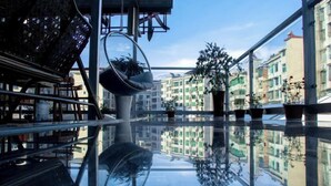 A modern interior with a glass floor reflecting the buildings outside, a hanging chair, and potted plants. - Mugen Town Residence (Zixing)