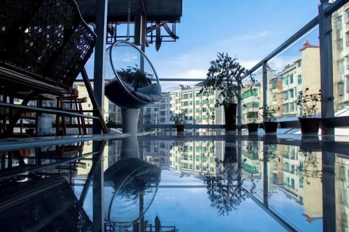 A modern interior with a glass floor reflecting the buildings outside, a hanging chair, and potted plants.
