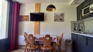 Ein Essbereich mit Tisch und Stühlen, ein an der Wand befestigter Fernseher sowie eine Küchenzeile mit Schränken und einer Mikrowelle.