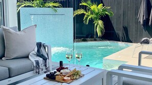 Un área junto a la piscina con un sofá, una mesa con aperitivos y copas de vino, y vista al estanque.