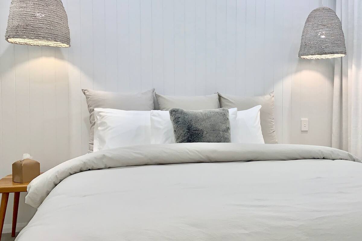 A neatly made bed with a white duvet and several pillows, including a fluffy one. There are two wall-mounted lamps with a woven design. A small wooden nightstand with a tissue box is visible to the left.