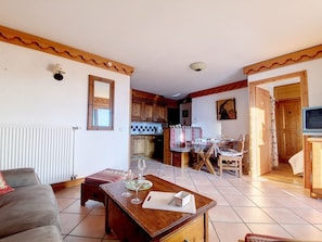 Sala de estar com sofá, mesa de jantar de madeira com cadeiras, cozinha com armários de madeira e televisão em suporte de madeira.