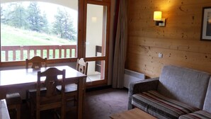 Een kamer met een houten tafel en stoelen, een bank, een raam met uitzicht op bomen en een wandlamp.