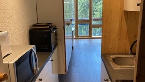 A compact kitchen with a microwave, toaster, and sink. A sliding door leads to a balcony with a view of trees.