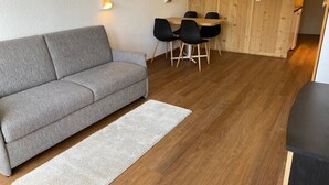 A modern living room with a grey sofa, a wooden dining table with chairs, and a light-colored rug on a wooden floor.