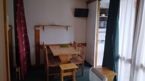 A compact living space with a wooden dining table, chairs, a small green bowl, a TV mounted on the wall, and a kitchenette visible through an open door. - Studio for 4, slopeside with balcony (Plagne Bellecôte)