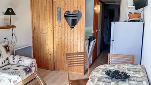 A compact room with a wooden wardrobe, a bed with patterned bedding, a small table with a decorative item, and a television mounted on the wall.