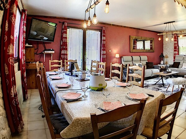 A dining area with a long table set for six, surrounded by wooden chairs. A television is mounted on the wall, and there are hanging light fixtures above the table.