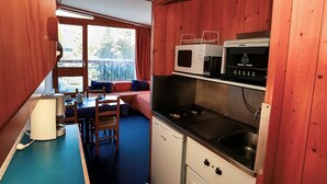 Una cocina compacta con microondas, cafetera y un lavamanos pequeño. Hay una zona de comedor con mesa y sillas, y un ventanal con vistas al exterior.