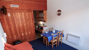 Una habitación pequeña con cama, mesa y sillas, una kitchenette y una pared blanca con una rejilla de ventilación.