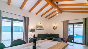 Una sala moderna con techo de madera, un sofá, una mesa de comedor con copas de vino y vista al mar a través de amplios ventanales.