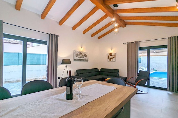 A modern living room with a wooden ceiling, a sofa, a dining table with wine glasses, and a view of the sea through large windows.