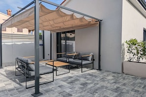 A covered outdoor seating area with two tables and four chairs, a pergola with a beige canopy, and a potted plant on the right.
