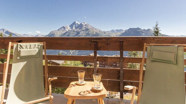 A wooden table with two chairs, a glass, and a plate, set against a mountain backdrop.