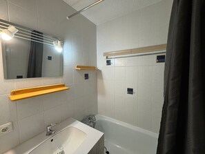Un baño con lavamanos blanco, espejo y bañera. Hay repisas de madera sobre el lavamanos y un perchero para toallas.