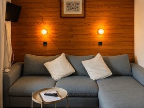 Una sala de estar moderna con un sofá gris, dos almohadas blancas, una mesita redonda con un control remoto y dos lámparas fijadas a la pared.