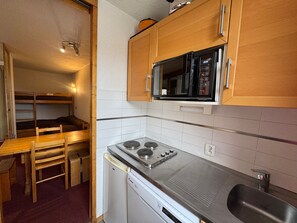 Cocina compacta con microondas, estufa y lavavajillas. Hay una mesa de madera y una cama litera en la zona de dormir contigua.