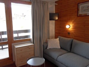Una habitación con pared de madera, un sofá gris, una pequeña mesa redonda, un televisor montado en la pared y una ventana con cortinas.