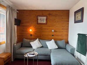 Una habitación con pared de madera, un sofá gris con cojines, una mesita con una revista, un televisor montado en la pared y un cuadro enmarcado sobre el sofá.