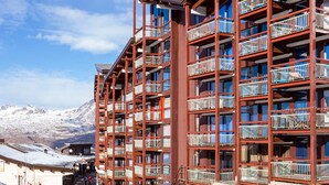 A multi-story hotel with balconies, a ski slope in the background, and a mountainous landscape.