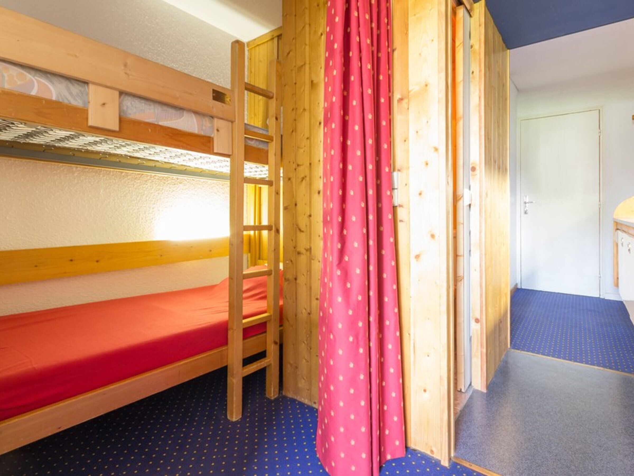 Een slaapzaal met stapelbedden, een houten trapje, een rood gordijn en een badkamer met een blauwe mat.