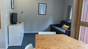 Um quarto com uma mesa de madeira, uma escrivaninha branca com um computador, um sofá cinzento com um travesseiro amarelo e um quadro na parede.