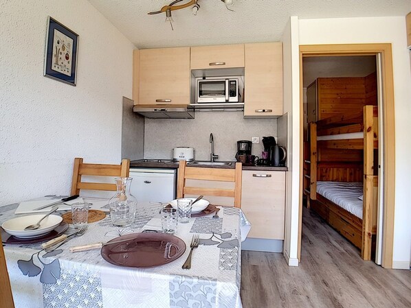 A compact kitchen with a microwave, stove, and coffee maker. There is a dining table set with plates, glasses, and cutlery. A bunk bed is visible in the adjacent room.