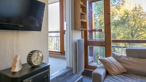 Una stanza accogliente con televisore a parete, una mensola in legno con oggetti decorativi, un divano con cuscini e un'ampia finestra che dà su alberi.