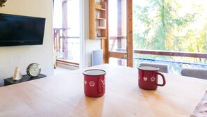 Una stanza con un tavolo di legno, due tazze rosse decorate con motivi bianchi, un televisore a schermo piatto, una sveglia e vista su alberi attraverso una finestra.