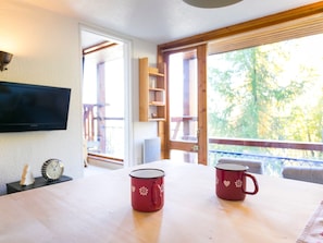 A room with a wooden table, two red mugs with white designs, a flat-screen TV, a clock, and a view of trees through a window.