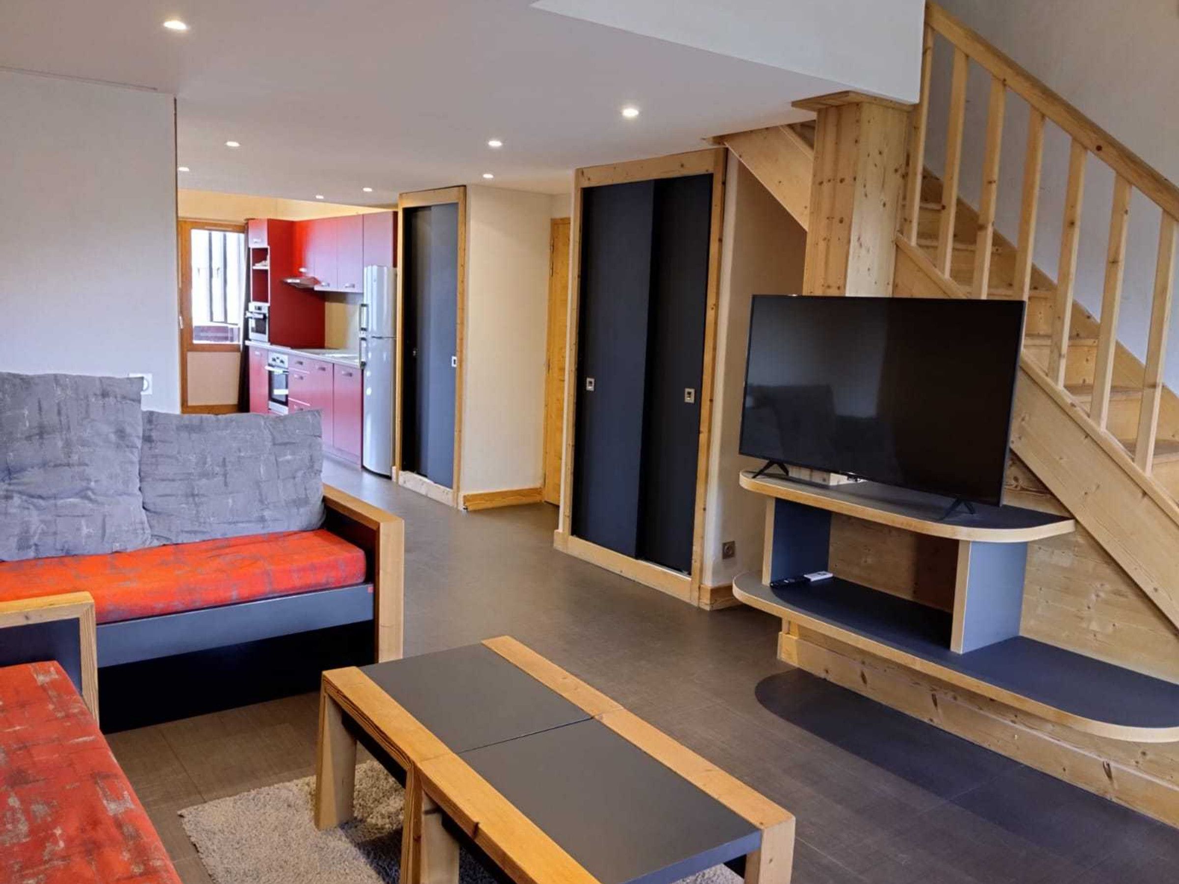Ein modernes Wohnzimmer mit einer Couch, einem Fernseher auf einem Holzschrank, einer Treppe und einem Küchenbereich im Hintergrund.