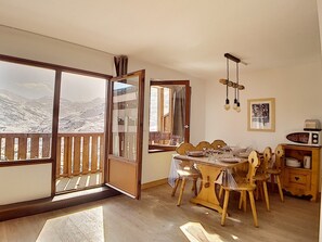 Een eetgedeelte met een tafel voor vier personen, houten stoelen, een houten kast met keukenapparatuur en uitzicht op besneeuwde bergen door een glazen deur.