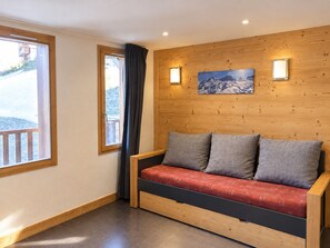 Ein Sofa mit Holzrahmen und grauen Kissen, eine Holzwand mit einem gerahmten Bild und ein Fenster mit Blick auf eine verschneite Landschaft.