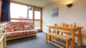 A cozy room with a wooden bench, a small table with kitchenware, a sofa with a checkered blanket, and a large window with curtains.